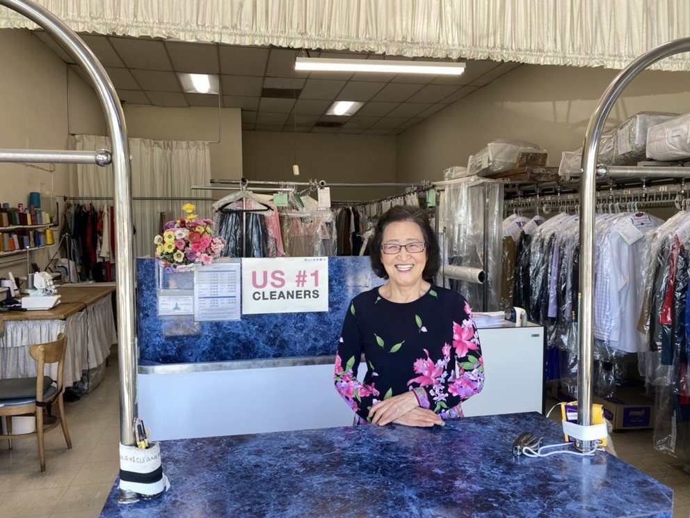 The shop owner standing behind the blue marble front counter, smiling, with rails of bagged clothes behind her.
